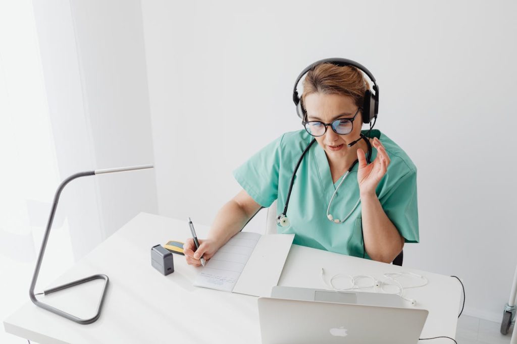 pexels-photo-7195445 Doctor in scrubs with headset engages in an online consultation while taking notes.